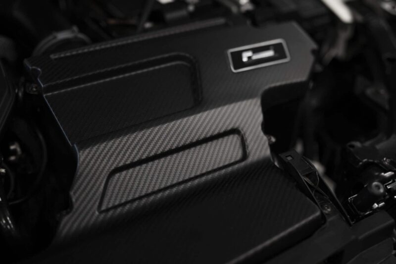Close-up of a carbon-fiber engine cover with a small rectangular badge and visible bolts in a dark automotive engine bay.