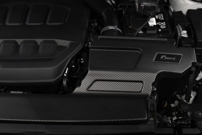 Close-up of a car engine bay with carbon-fiber panels and a visible battery on the right side.