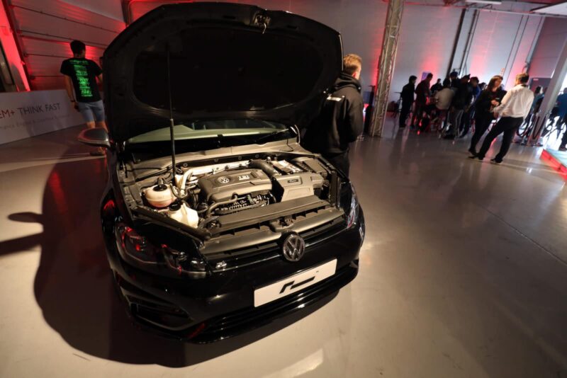 Black VW car with hood open on display at an indoor event, engine visible and people standing in the background.