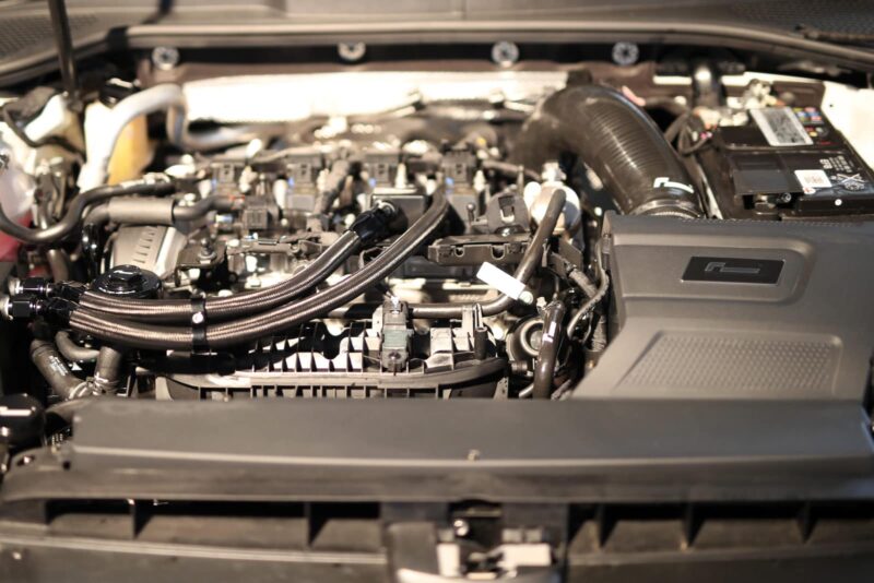 Car engine bay with hoses, wiring, and a large air intake on the right under the hood of a car