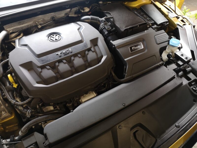 Engine bay of a Volkswagen with a prominent TSI engine cover and VW emblem, showing air intake hose and surrounding components.
