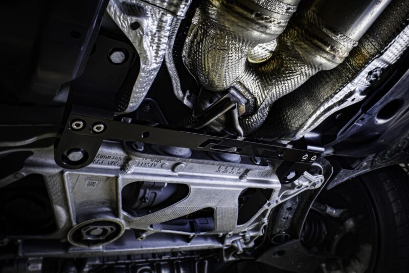 racingline-front-subframe-brace-9_51826108513_o-scaled-2jpg - OPSHOLDERS Underside of a vehicle showing exhaust pipes, heat shields, and metal chassis in a dimly lit area.