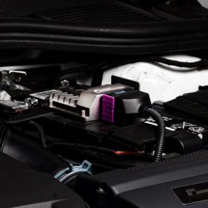 Under-the-hood shot of a car battery with a jump-start/charging device connected to the terminal, featuring a purple clamp.