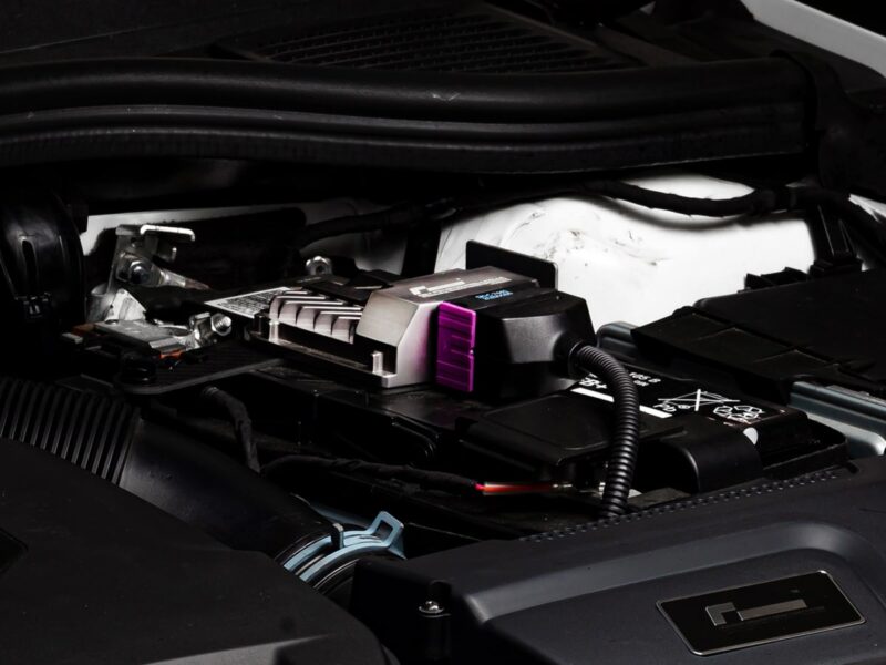 Under-the-hood shot of a car battery with a jump-start/charging device connected to the terminal, featuring a purple clamp.