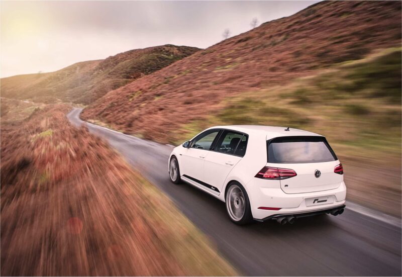 White SUV driving along a winding hillside road at dusk, with brown-purple heather and blurred foreground suggesting speed.