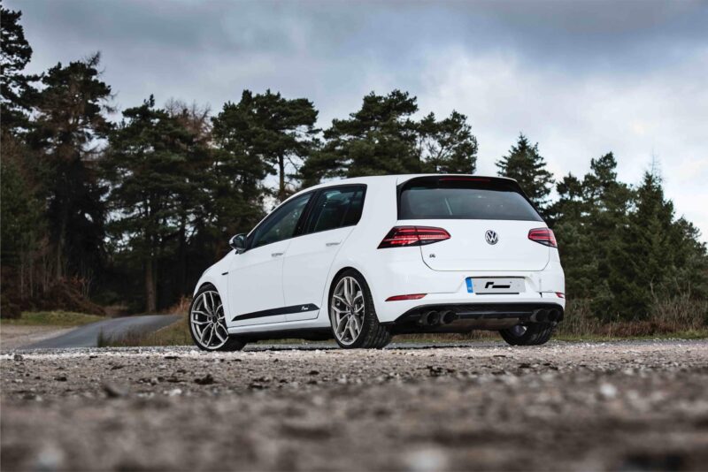 Rear view of a white Volkswagen hatchback parked on a gravel road with tall evergreen trees and a cloudy sky in the background.