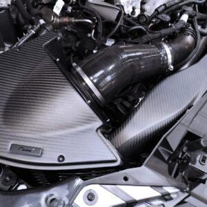 vwr1241s4_49398903913_o-1jpg - OPSHOLDERS Close-up of a car engine bay showing a carbon fiber panel and a large curved air intake duct with metal clamps.