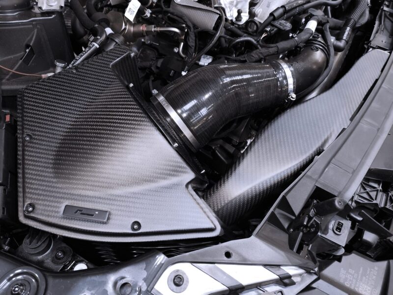 vwr1241s4_49398903913_o-1jpg - OPSHOLDERS Close-up of a car engine bay showing a carbon fiber panel and a large curved air intake duct with metal clamps.