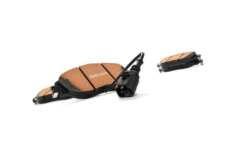 Close-up of tan friction-material brake pads with black backing plates and a sensor wire, laid on a white background.
