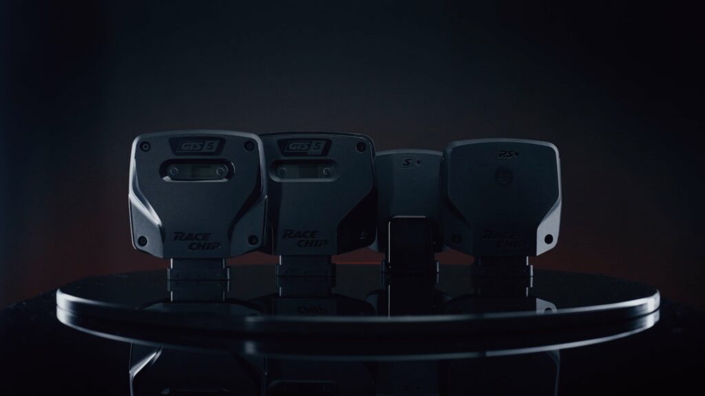Four dark devices with branding (GTS5 and Race Chip) lined up on a reflective circular table in a dim studio backdrop, showing their backs.