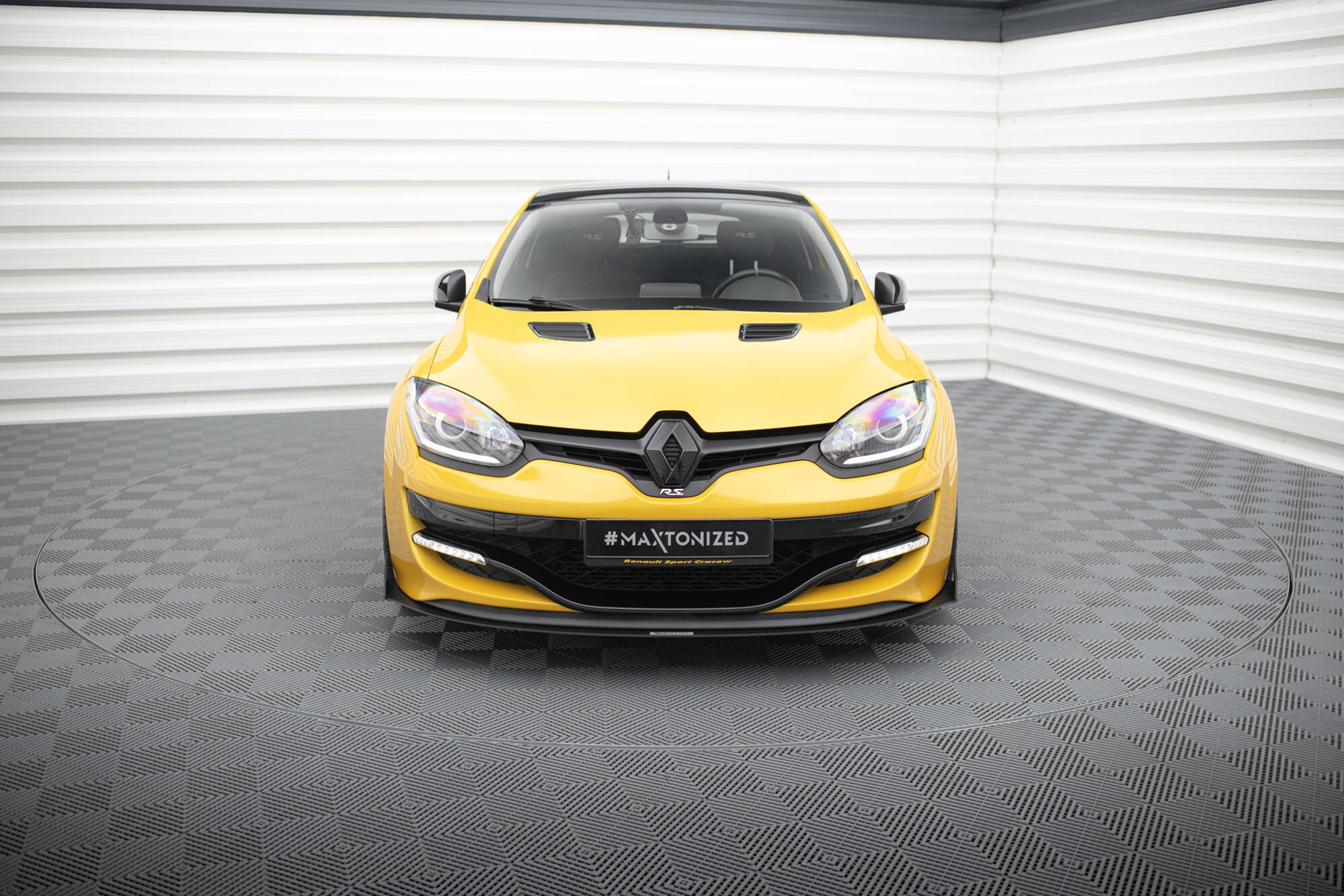 pol_po_Bonnet-Vents-459_1jpg - OPSHOLDERS Front view of a bright yellow Renault car parked indoors against white corrugated walls, showing the grille, headlights, and a black license plate reading #MAXTONIZED.