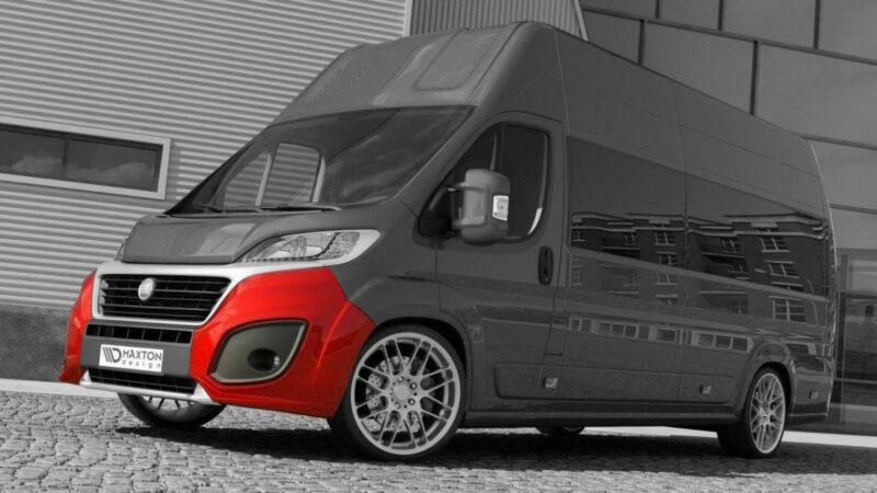 Black and gray delivery van with a bright red front bumper parked on a cobblestone surface, reflecting a nearby building in the side panels.