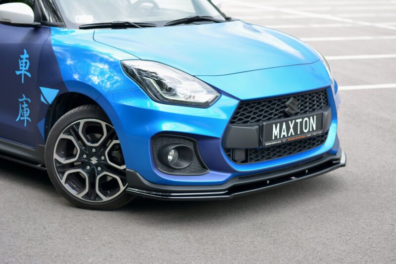 pol_po_FRONT-SPLITTER-V-1-suitable-for-Suzuki-SWIFT-6-SPORT-8268_2jpg - OPSHOLDERS Close-up of a bright blue car’s front-left section, showing the headlight, grille, and a 'MAXTON' license plate in a parking lot.