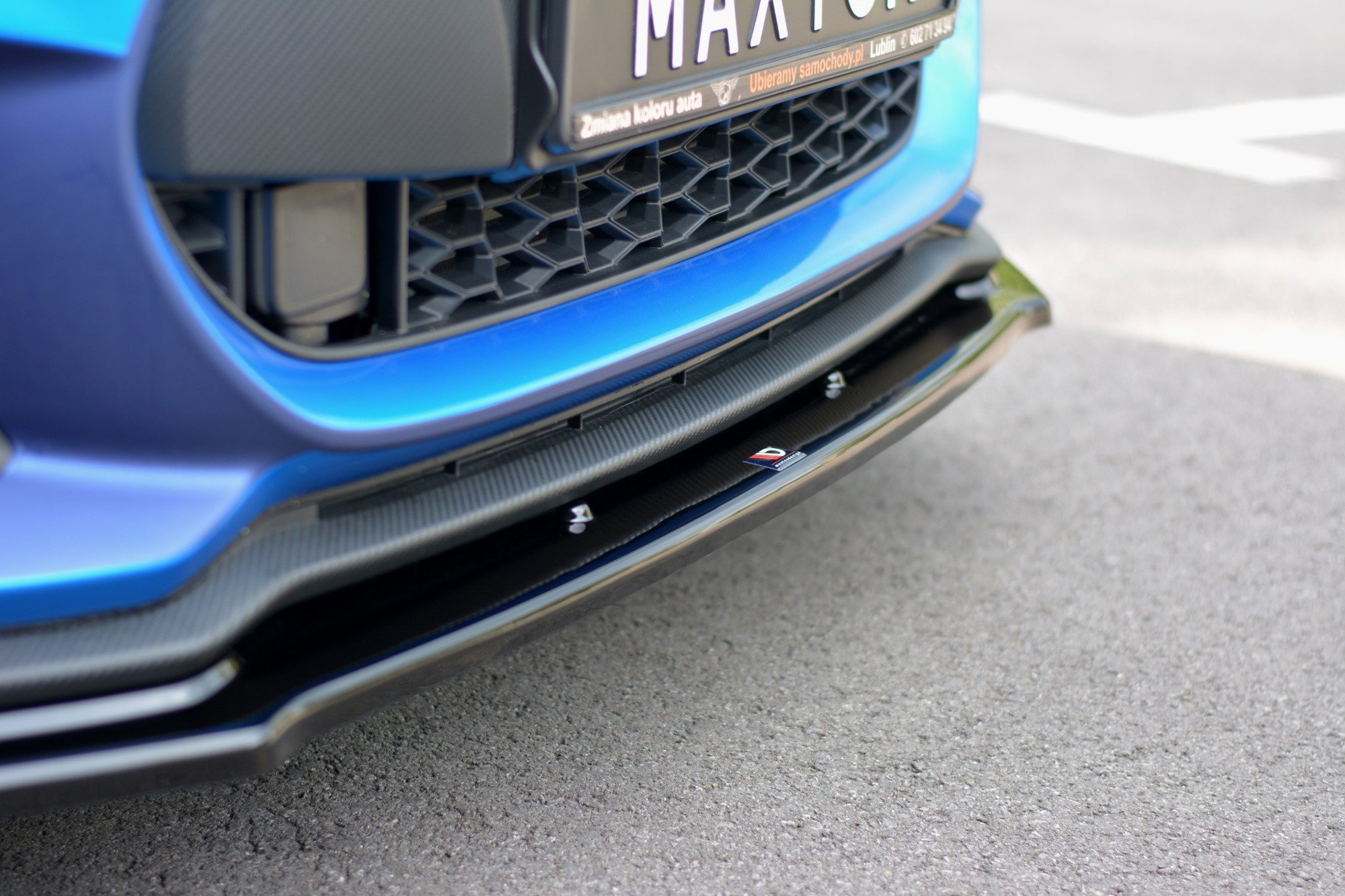 pol_po_FRONT-SPLITTER-V-1-suitable-for-Suzuki-SWIFT-6-SPORT-8268_4jpg - OPSHOLDERS Close-up of the front of a blue car showing a honeycomb grille and carbon-fiber lip under the bumper, with part of the license plate visible.