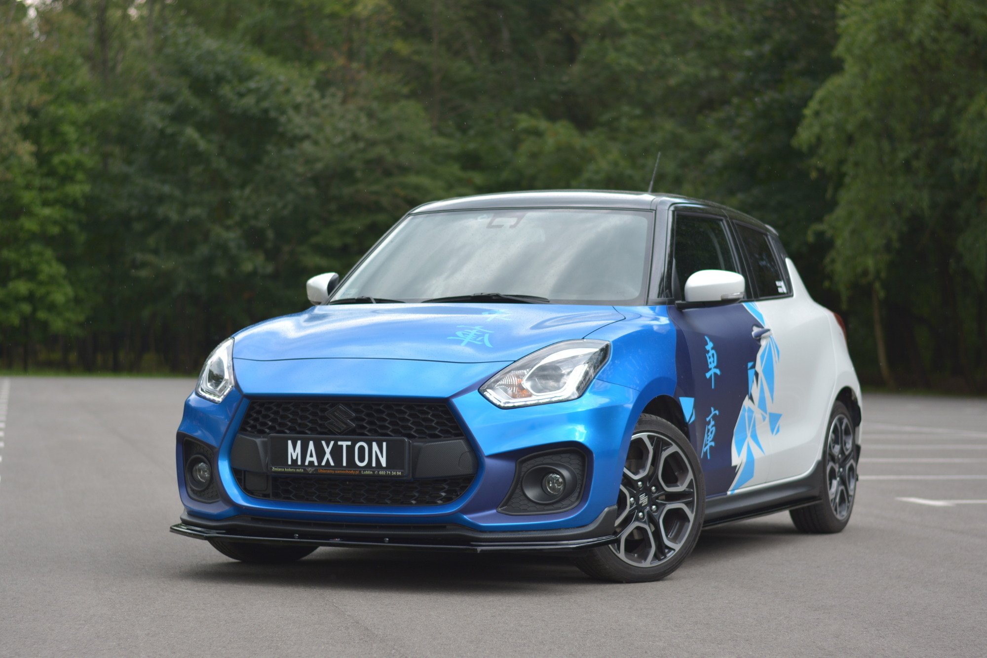 pol_po_FRONT-SPLITTER-V-1-suitable-for-Suzuki-SWIFT-6-SPORT-8268_7jpg - OPSHOLDERS Colorful blue and white compact car with racing decals, front view in a parking lot, license plate reads MAXTON, green trees in the background