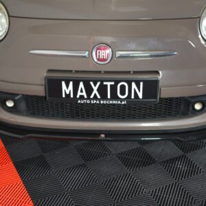 Front view of a brown Fiat car showing its circular headlights, badge, and the 'MAXTON' license plate on a perforated front bumper plate holder on a showroom floor.