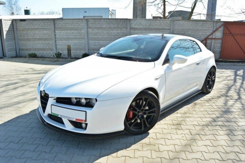 White sports coupe parked on a paved driveway, front-left angle, black multi-spoke wheels, red brake calipers, brick/stone fence in background.