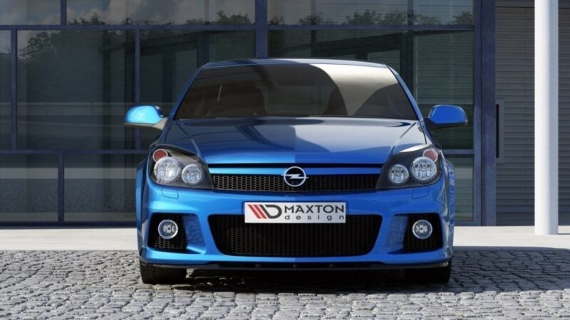 Front view of a blue sports hatchback with a Maxton design plate, parked on a cobblestone driveway in front of a glass-front building.