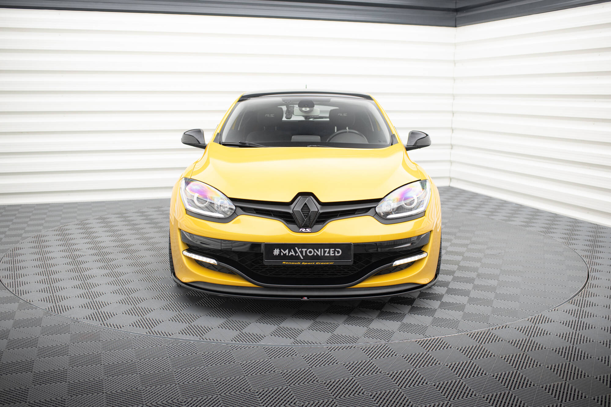 pol_po_FRONT-SPLITTER-suitable-for-Renault-MEGANE-3-RS-v-1-872_4jpg - OPSHOLDERS Front view of a bright yellow Renault car in a showroom, showing the grille and headlights, with '#MAXTONIZED' on the license plate area.