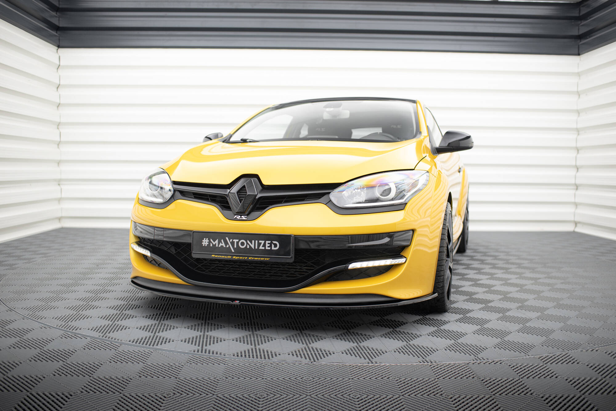 pol_po_FRONT-SPLITTER-suitable-for-Renault-MEGANE-3-RS-v-1-872_5jpg - OPSHOLDERS Yellow Renault car facing forward in a showroom, close-up of front with prominent Renault badge and black grille, '#MAXTONIZED' plate visible