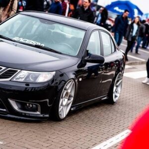 Black sedan with lowered suspension and large silver wheels at a car show, surrounded by onlookers.