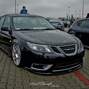 A modified black sedan with a lowered stance and large silver multi‑spoke wheels, parked at an outdoor car show with people in the background.