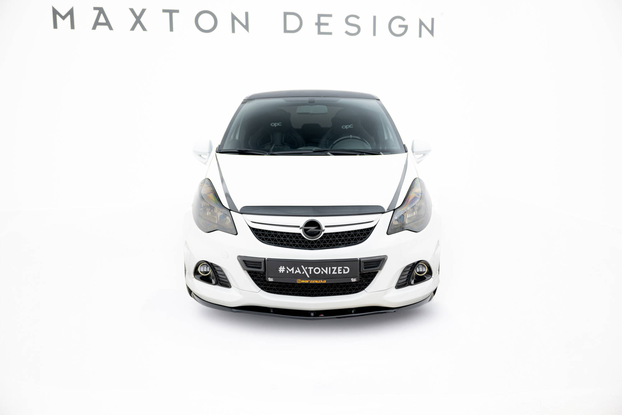 pol_po_Front-Splitter-suitable-for-Opel-Corsa-D-OPC-VXR-840_4jpg - OPSHOLDERS Front view of a white car in a studio with a black grille and Opel badge, displaying the '#MAXTONIZED' plate and 'MAXTON DESIGN' text above.