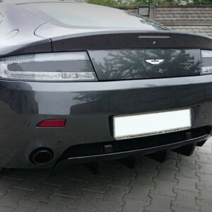 Rear view of a dark gray Aston Martin sports car on a paved driveway, showing taillights, badge, and a prominent rear diffuser with dual exhausts.