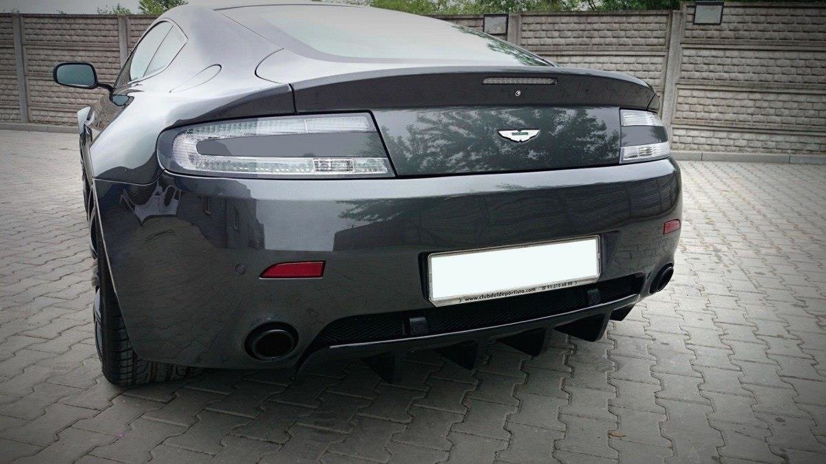 Rear view of a dark gray Aston Martin sports car on a paved driveway, showing taillights, badge, and a prominent rear diffuser with dual exhausts.