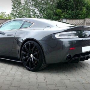 Gray Aston Martin sports coupe, rear three-quarter view, large black multi-spoke wheels on a paved driveway.