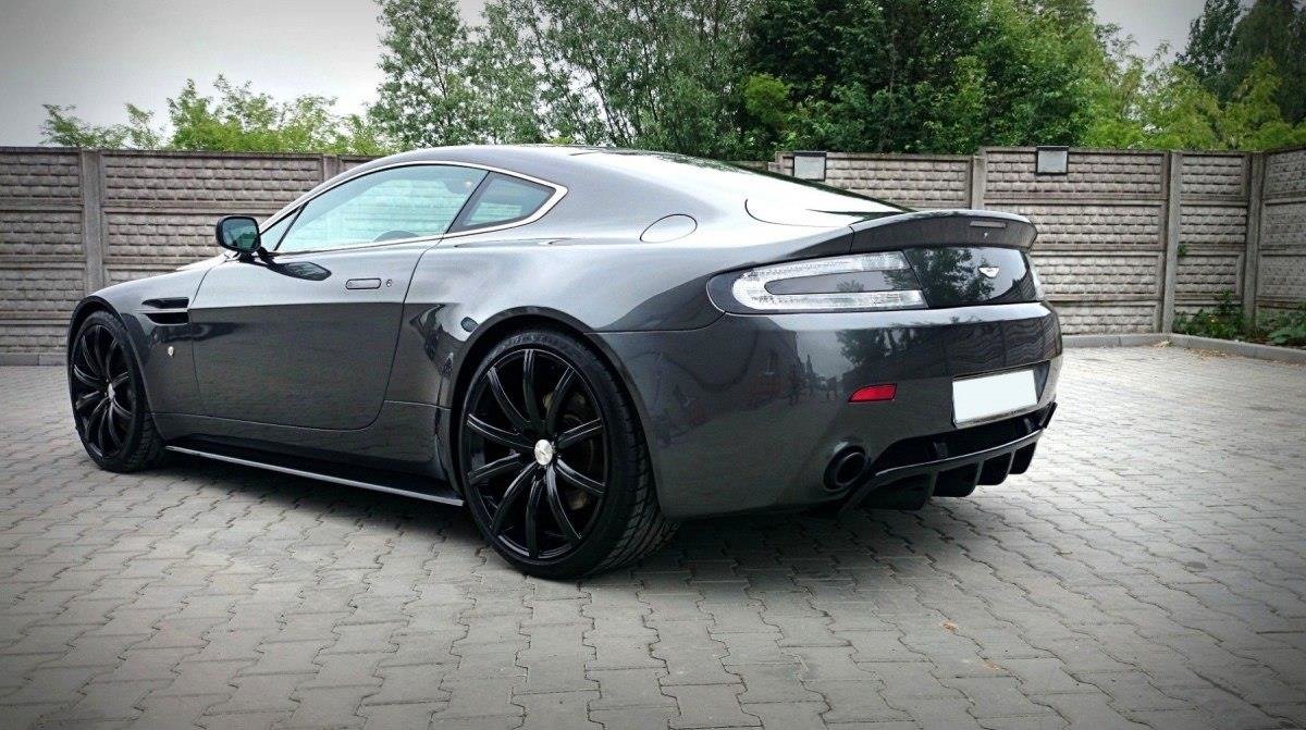 Gray Aston Martin sports coupe, rear three-quarter view, large black multi-spoke wheels on a paved driveway.