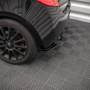 Rear section of a dark car, showing the back wheel and exhaust with a glossy body in a showroom with a patterned floor surface.