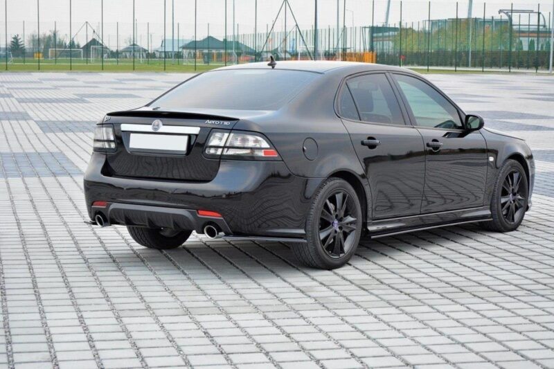Rear three-quarter view of a black four-door sedan parked on a patterned paved area with a sports field in the background.