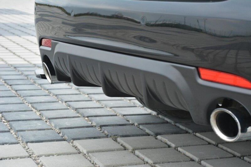 Close-up of the rear bumper of a dark car with two chrome exhaust tips and a sculpted diffuser, parked on a paved surface.