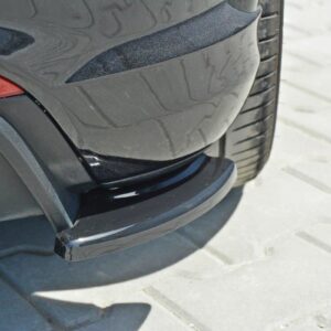 Close-up of a dark car's rear bumper with a protruding black protective lip, parked on a paved surface with a visible tire nearby