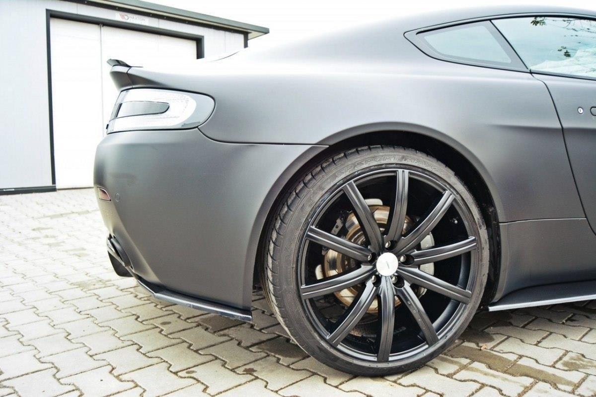 Rear-side view of a gray sports car with a large black multi-spoke alloy wheel and visible brake disc on a paved driveway/lot.