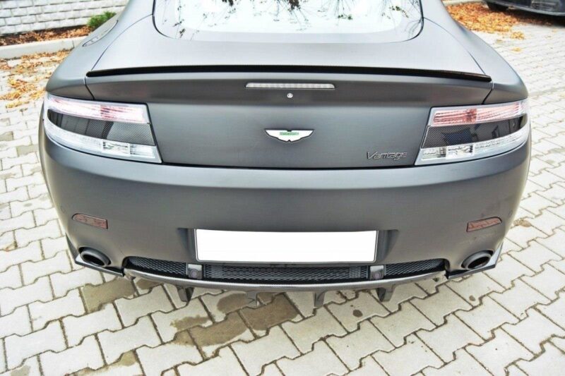Rear view of a matte gray Aston Martin Vantage, showing taillights, emblem, and rear diffuser on a brick-paved surface.