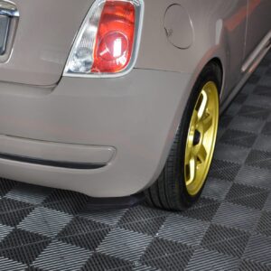 Rear of a gray car showing taillight and license plate, with a yellow wheel rim visible on a checkered floor surface.