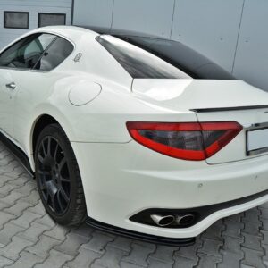 Rear three-quarter view of a white Maserati sports car parked on a brick driveway beside a gray building.