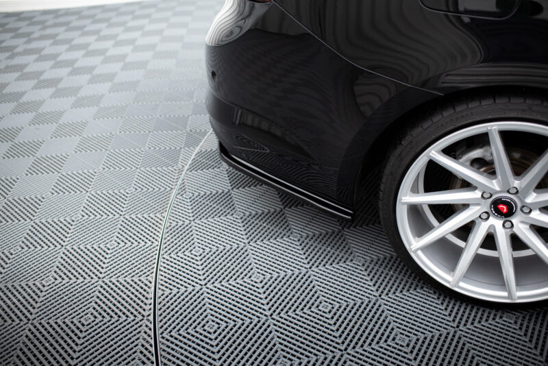 pol_po_Rear-Side-Splitters-suitable-for-Renault-Talisman-1143_4jpg - OPSHOLDERS Close-up of a black car's rear quarter, halogen wheel and tire visible on a patterned metal floor with a white cord lying on the ground.