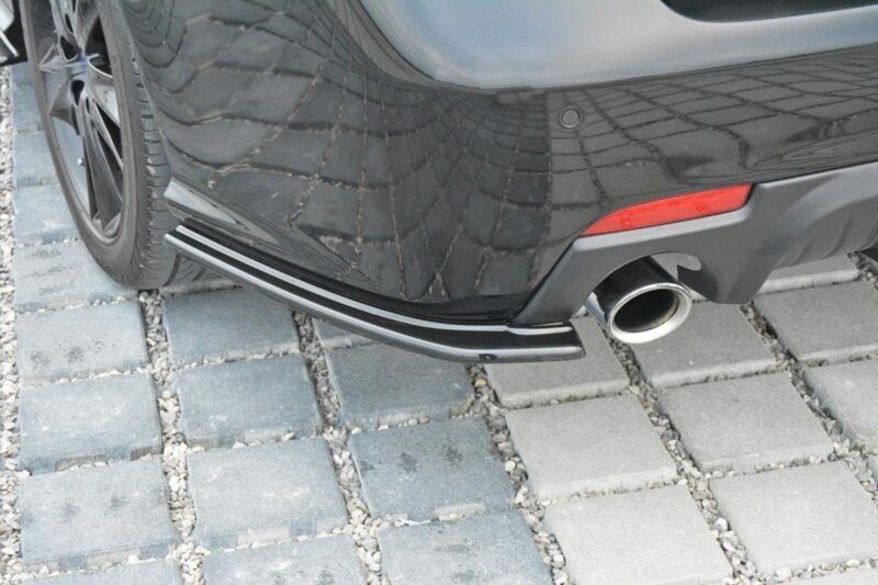 Rear view of a black car showing a chrome exhaust tip and bumper on a gray paving stone surface.