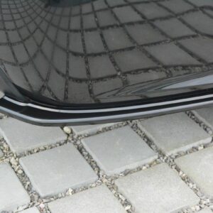 Close-up of a black car's rear bumper and exhaust pipe, parked on a stone-paved driveway with square pavers.