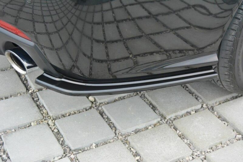 Close-up of a black car's rear bumper and exhaust pipe, parked on a stone-paved driveway with square pavers.