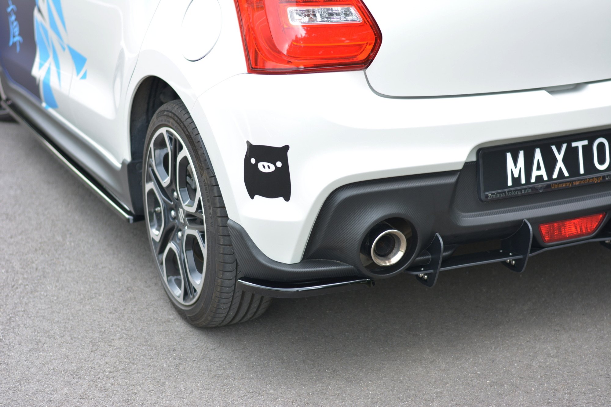 pol_po_Rear-Side-Splitters-suitable-for-Suzuki-Swift-Mk6-8260_1jpg - OPSHOLDERS White car rear featuring a black pig sticker on the rear fender near the wheel and a bold rear bumper with a visible exhaust.