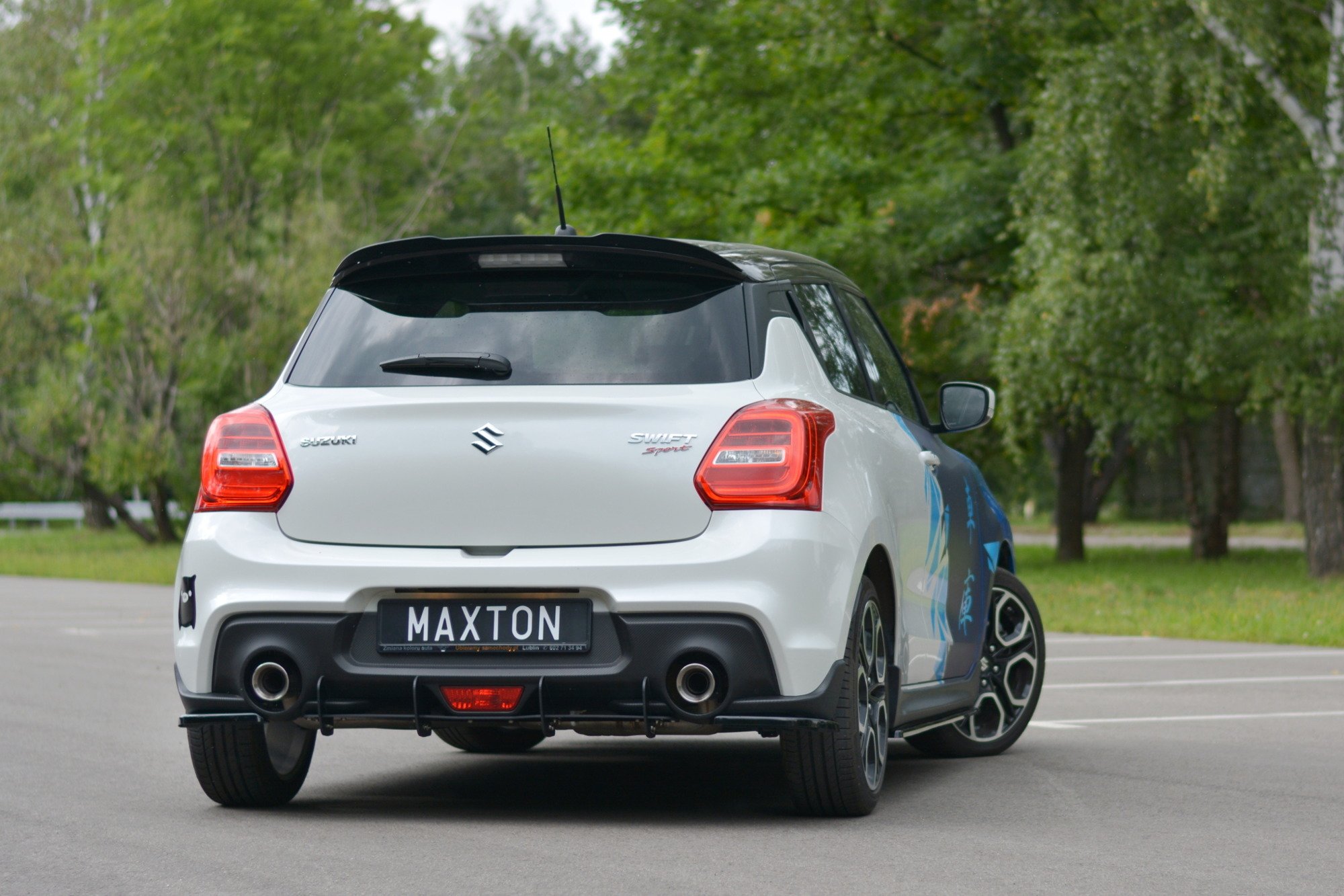 pol_po_Rear-Side-Splitters-suitable-for-Suzuki-Swift-Mk6-8260_4jpg - OPSHOLDERS Rear view of a silver Suzuki Swift Sport with a black spoiler and Maxton license plate, parked on pavement with trees in the background.
