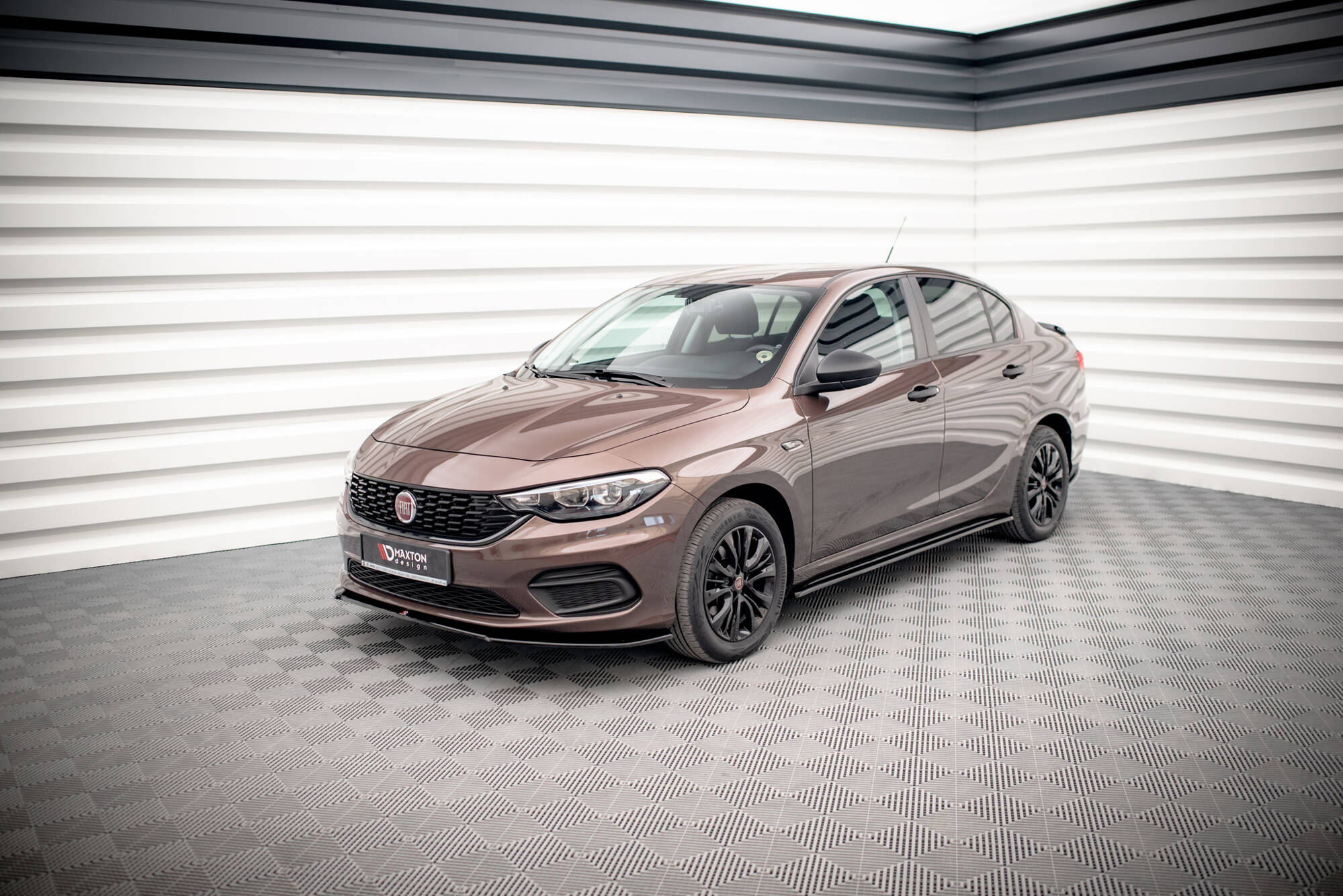 Brown Fiat sedan photographed in a bright, minimalist showroom with white paneled walls and a checkered floor, shown from the front-right.