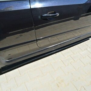 Black car with a lower side skirt and alloy wheels parked on a light-paved surface, reflecting the surroundings on its door panel.