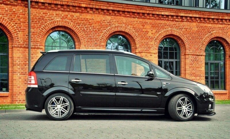 pol_po_SIDE-SKIRTS-DIFFUSERS-suitable-for-Opel-ZAFIRA-B-OPC-254_5jpg - OPSHOLDERS Black MPV parked on a paved lot in front of a red brick building with arched windows.
