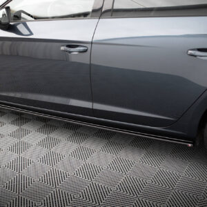 Close-up side view of a gray sedan showing the front and rear doors with alloy wheels on a patterned floor surface, showroom-style lighting.