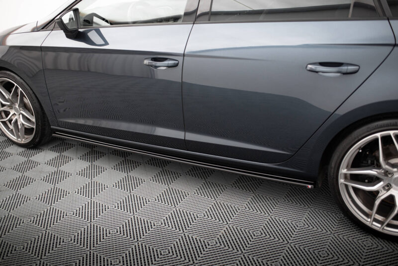 Close-up side view of a gray sedan showing the front and rear doors with alloy wheels on a patterned floor surface, showroom-style lighting.